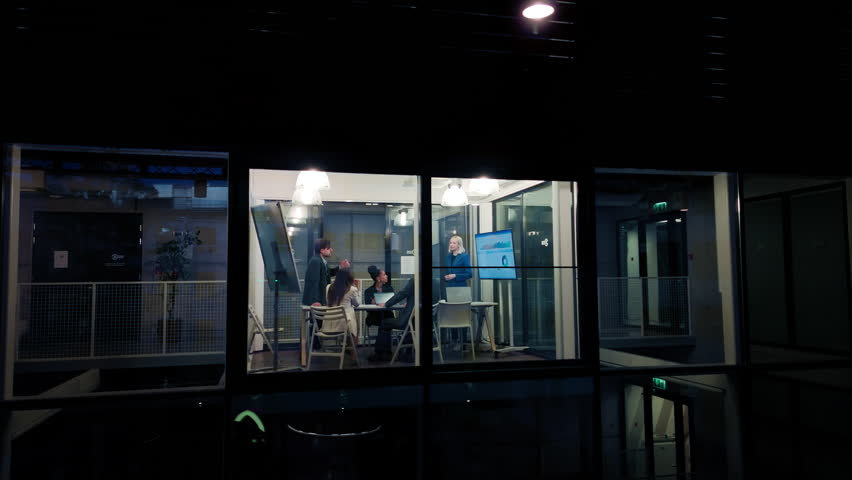 Group of Corporate Workers Analyzing Results From a Marketing Campaign. Project Manager Presenting Data Chart on Screen, Employees Having a Conversation in a Meeting Room. View from Outside the Window