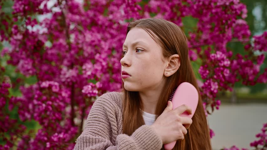 A teenage girl with long, brown hair stands in front of a backdrop of pink flowers. She is wearing a light pink sweater and looking directly at the camera.
