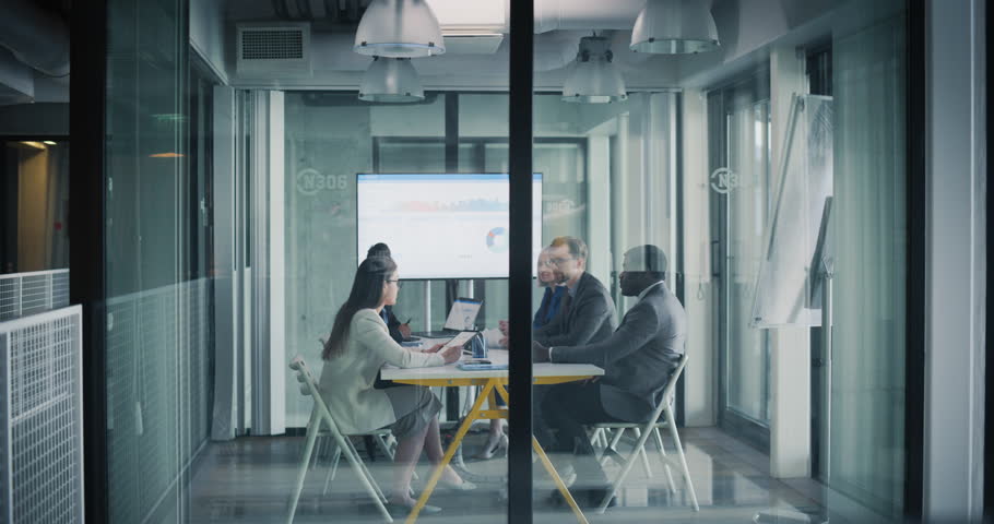 Group of Corporate Workers Analyzing Results From an Advertising Campaign. Marketing Team Presenting Data Charts on Screen, Employees Having Conversations in a Glass Meeting Room