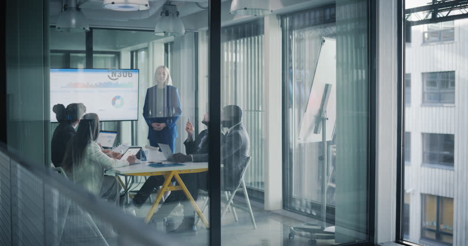 Group of Corporate Workers Analyzing Results From a Marketing Campaign. Project Manager Presenting Data Chart on Screen, Employees Having a Conversation in a Meeting Area. View From Outside the Room