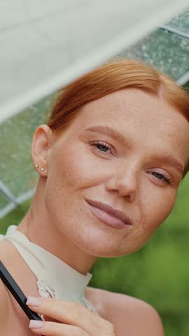 A woman relaxes while holding a transparent umbrella, enjoying the rain amidst vibrant greenery.