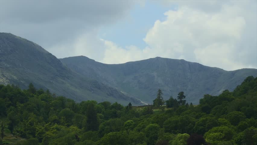 Clouds blowing from behind mountains as patches of sunlight race over mountains and trees. Time lapse. Lake District, Cumbria, UK