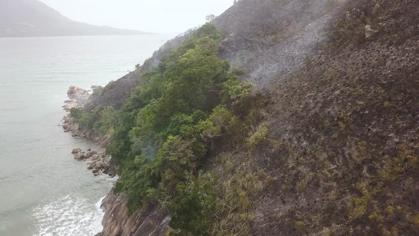 Smoke rising from recently burned vegetation on seaside mountain. Aerial backwards