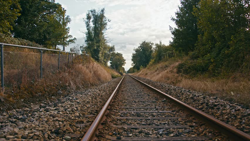 The camera pans up from the ground, revealing railway tracks stretching into the distance. The view highlights the rails as they lead through a picturesque landscape, emphasizing depth and journey.