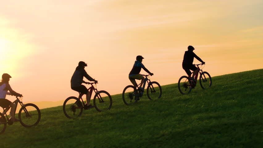 AERIAL, SILHOUETTE, LENS FLARE: Stunning sunset with group of mountain bikers riding up the grassy hill. Picturesque bike ride through the green, hilly Slovenian countryside in beautiful golden light.