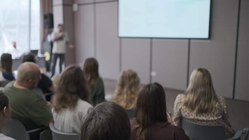 Group of people attentively listens to a presentation in a modern conference room. The speaker is presenting at the front with a large screen displaying visuals.