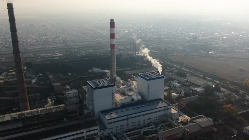 A wide-angle view of a large industrial power plant with tall chimneys emitting smoke, surrounded by a densely packed urban area under a foggy sky
