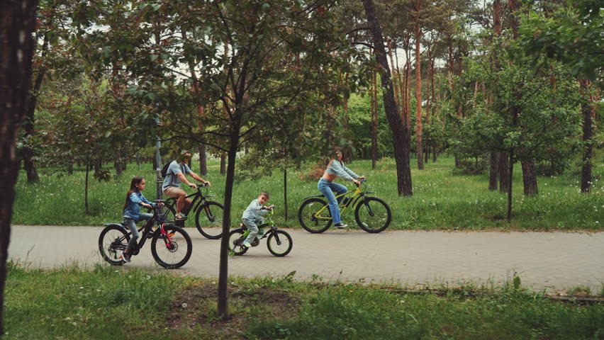 Family of four biking together on park path surrounded by lush greenery and trees. Concept of family bonding outdoor adventure and healthy lifestyle
