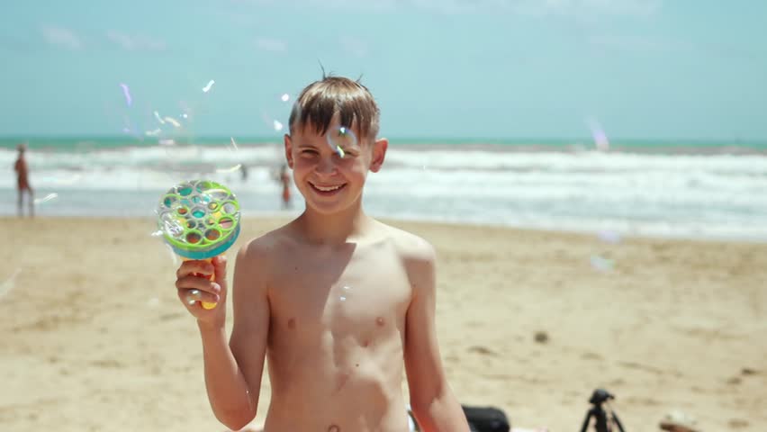 boy having fun on sunny beach, playing with colorful bubble gun and making big soap bubbles. Slow motion