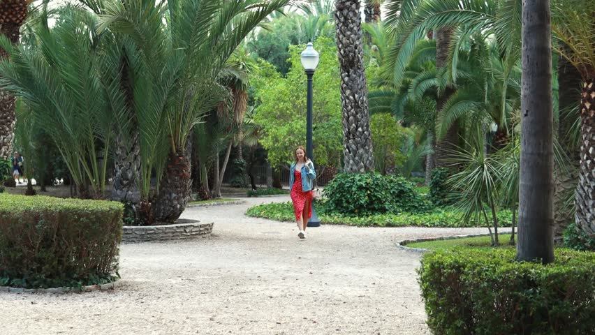 Young woman wearing red dress and blue jacket is walking in park. Girl smiling and enjoy sunny day.  slow motion