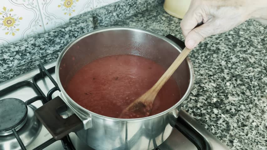 Close-up of elderly woman's hands stirring natural homemade tomato sauce in a stainless steel pot using a wooden spoon. Traditional cooking with fresh ingredients, showcasing authentic Italian culinar