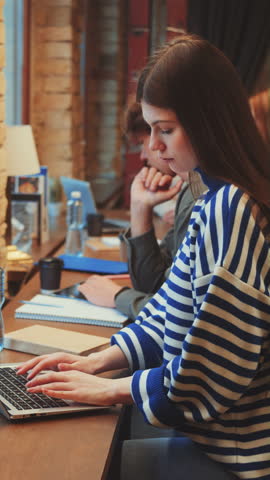 Vertical screen: Students sit at window-side desks in library, focused on laptops and notebooks. Cozy atmosphere with exposed brick and plants. Concept of dedicated study and academic productivity