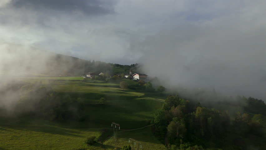 Aerial view of a quaint village nestled in lush green hills, partially covered in morning fog, creating a serene and peaceful atmosphere.