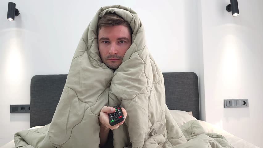 White man wrapped in blanket looks sick or tired holding remote control while sitting in bed as he listlessly flips through TV channels