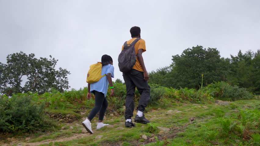 A man and a woman are walking up a hill. The man is wearing a yellow shirt and the woman is wearing a blue shirt. They both have backpacks on. The scene is peaceful and serene