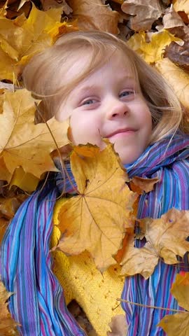 Little girl with light hair and bright scarf rests in golden leaves. Dressed in yellow, her joyful smile radiates warmth, embodying spirit of autumn and simple joys of nature