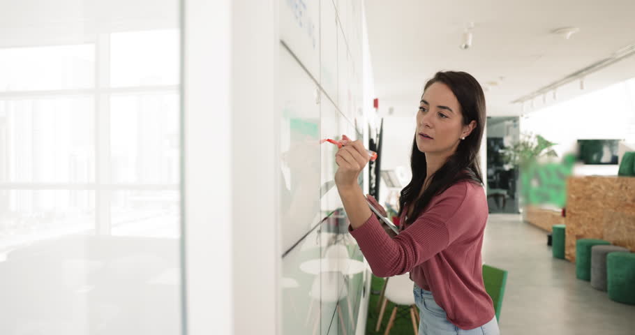 Young woman writing information on whiteboard, outlining project timelines, key points, listing topics to be discussed in upcoming meeting or seminar, jotting down ideas. Planning, goals, objectives