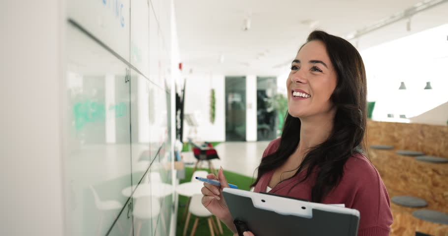 Young female, office employee holds folder prepare agenda, writing notes while stand at whiteboard with written info, check to-do lists, carefully reviewing important information. Workflow, planning