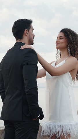 Portrait of happy man in suit and woman in white short dress and curly flying hair. Action. Lovely couple standing on a pier with the calm sea and cloudy sky on the background.