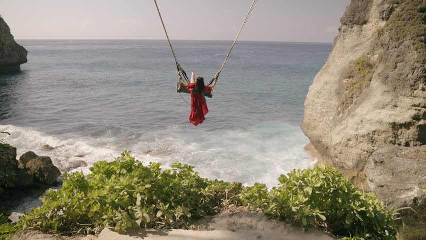 Nusa penida island, Bali, indonesia. 4K video Young woman red dress Play on the swings at Diamond beach, beutiful diamond beach,Nusa Penida island. Popular travel destination on Bali, indonesia.