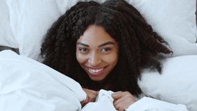 Happy playful african american woman peeking out from her blanket in bed and smiling to camera, top view, close up - Powered by Shutterstock - Get 15% off with code: PIKWIZARD15