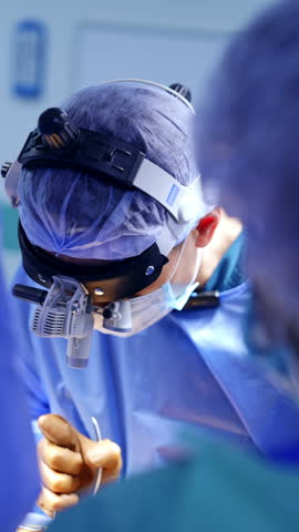 Vinnitsa, Ukraine, 22 June 2024: Portrait of a doctor wearing surgical loupes, cap and mask performing surgery. Top view. Surgeon gives the used tool to the assistant. Vertical video.