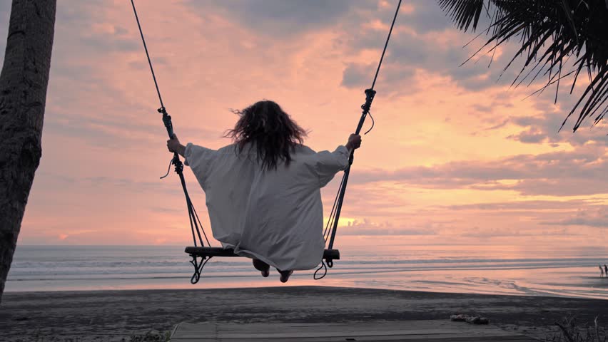 Woman Relax on Beach by Sea at Sunset. One Young Female Swings on Swing on Summer Evening by Ocean. Concept Happy Girl Rest on Tropical Island Sea Sunshine Background. Back View Alone Person Outdoor
