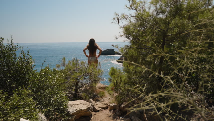 A Woman in a Swimsuit Standing on a Cliff Edge, Tossing Her Hair in Slow Motion. The Scenic Ocean View and Surrounding Pine Trees Create a Breathtaking Backdrop, Capturing a Moment of Freedom and Natu