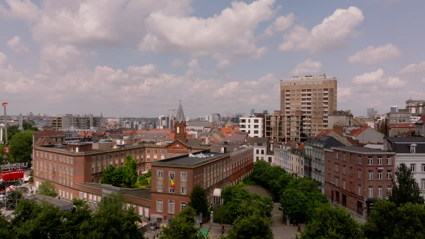 Birds eye view of the Brussels skyline pans across rooftops, a church steeple, and apartment buildings on a beautiful day with blue skies and fluffy white clouds