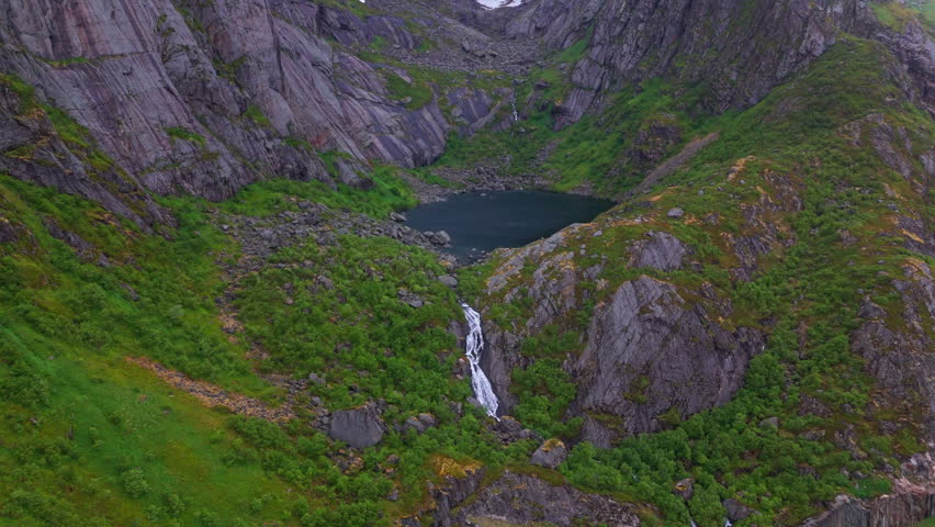 Olkonvatnet near Reine in Lofoten Norway. Small snow melting overflow pond on a mountain range, with a waterfall streaming down towards the sea. Mountain peaks on all sides, green vegetation on ground