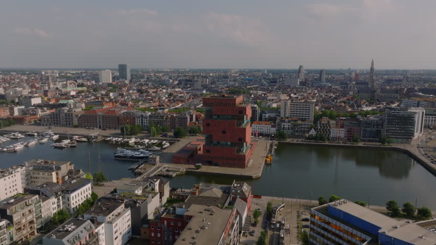 Aerial views highlight the MAS Museum aan de Stroom and the lively city of Antwerp. Boats glide along the Scheldt River while cars navigate busy streets beneath a clear blue sky