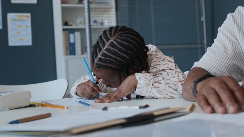 African American preschool girl colouring drawing with blue pencil during pediatric appointment at doctors office with her father