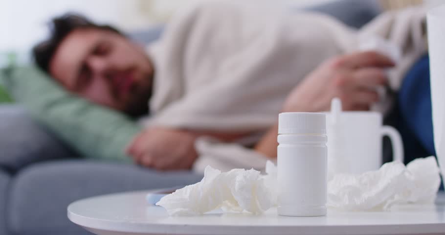 Sick young man lying on sofa at home with seasonal flu or cold feeling unhealthy with influenza and runny nose. Male person suffering from cough with a pills and napkins on table. 4k video.