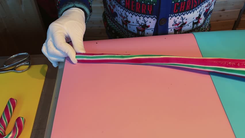 A person making Christmas sugar sticks cutting a ribbon with scissors on a pink table. The ribbon is striped and has a Christmas theme