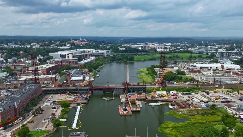 An aerial motion time lapse of the Norwalk Railroad Bridge on a cloudy day. There is construction around the bridge. The drone camera dolly in over the river towards the bridge and construction.