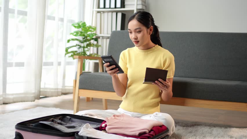 Young woman is checking her passport and using a smartphone while packing her luggage in her living room