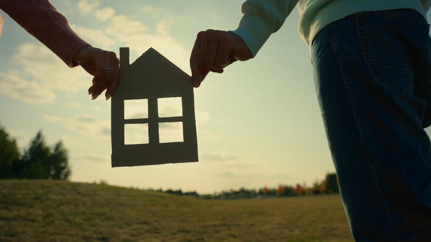 Happy Caucasian family woman man male female hands holding paper house outside sunset sun village nature field. Couple hold home model dream buying real estate investment mortgage bank loan insurance