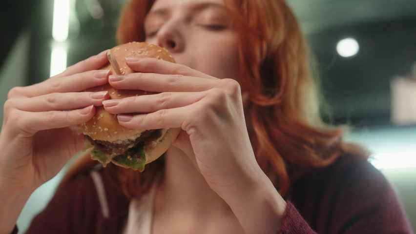 Happy hungry Caucasian woman female girl client customer eat chew tasty burger eating delicious juicy hamburger enjoy chewing cheeseburger indoors kitchen room. Fast food delivery unhealthy nutrition