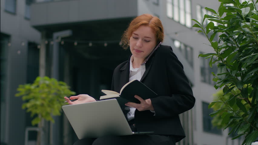 Busy successful Caucasian businesswoman ginger female girl woman sitting laptop notebook taking notes writing pen talking mobile phone cellphone call explain discuss business outside city urban street