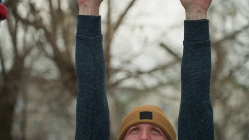 Close-up of a coach working out on red iron bars, visibly tired as he moves across the iron bar drops down, the blurred background features bare trees
