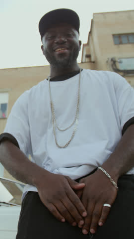 Vertical low angle shot of cool Black male rapper performing hip hop song on camera while leaning on car outdoors in back alley