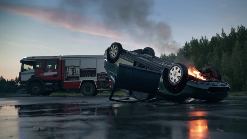 Dramatic Traffic Accident With a Rolled, Burning Compact Vehicle Emitting Thick Smoke. Fire Trucks Arrive on Scene with Emergency Signals. Nobody in Sight, Just the Crashed Car on the Road