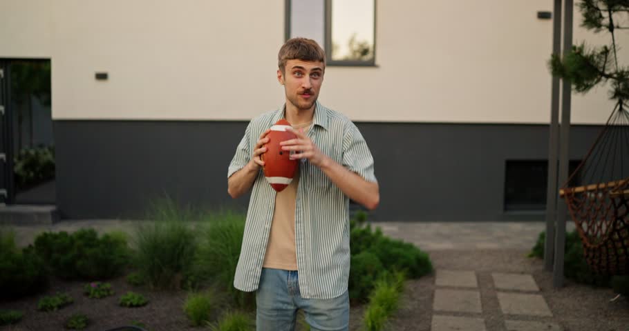 Happy blond guy with a mustache throws an American football ball to his friend while relaxing at a table in the backyard of a house