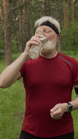 Vertical shot of senior man drinking water, setting up fitness tracker and running on forest path while training outdoors in nature on summer day