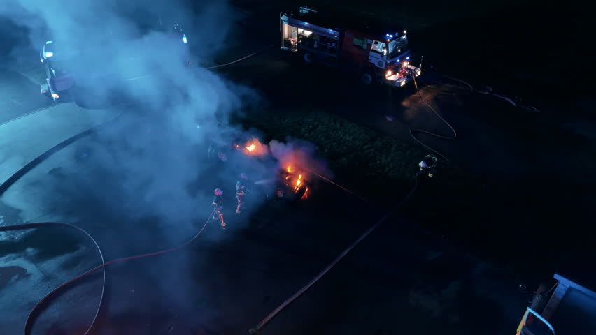 Aerial Drone View of Nighttime Car Accident Scene. Firefighters Combat Intense Vehicle Fire while Paramedics Take an Injured Person to the Emergency Ambulance on a Stretcher