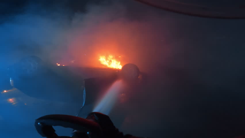 Intense POV Footage of a Firefighter Battling a Raging Fire on Rolled Car. Water Streams From a High Pressure Hose, Illuminated by an Explosion and Flames. Tragic Traffic Accident on a Motorway