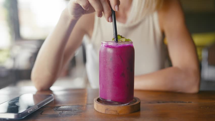 Young adult woman tourist enjoying exotic cocktails in Thai cafe in summer day. Closeup of cup with purple fruit smoothie on wooden table, lady stirring drink by plastic cocktail straw, relaxation