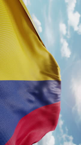 Wavy flag of Colombia blowing in the wind in slow motion. Waving colorful Colombian flag team symbol abstract vertical background. Blue sky with clouds. World countries flying flags concept