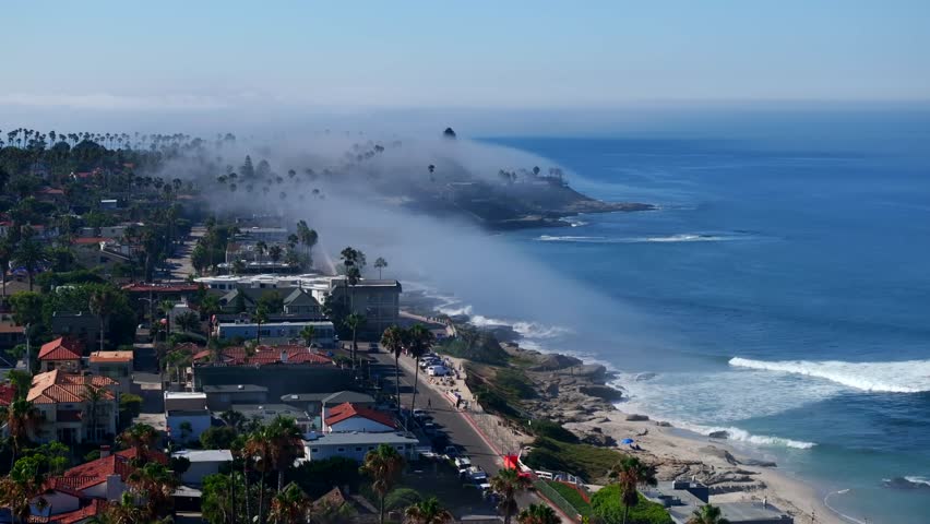 Aerial view of La Jolla coast with marine layer creating low flying fog clouds on an otherwise sunny day with blue sky in San Diego California with Pacific Ocean waves crashing along the shore