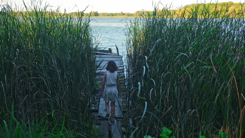 Cute Young Caucasian Girl Walking on Old Wooden Pier Through Reed Bed Rushes at Serene Lake Shore